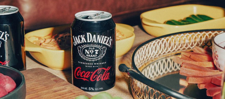 Image of the Jack Daniel's and Coca-Cola can sitting on a table at a party surrounded by snacks and vegetables in yellow serving dishes.