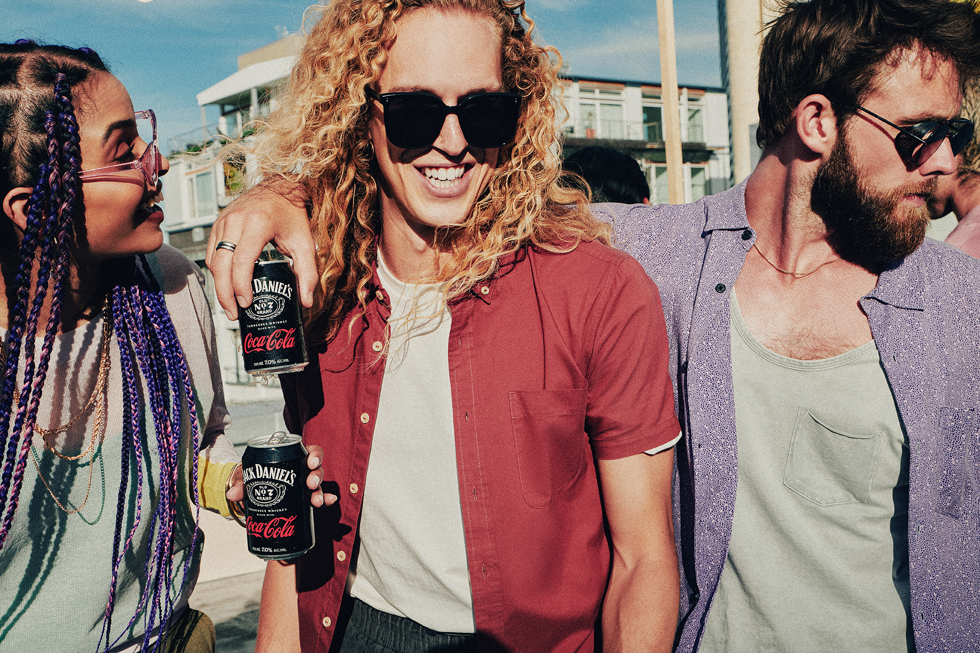 Image of a person in a red button up shirt with a man standing next to them to the right in a purple button up shirt with his arm around them holding a Jack Daniel's and Coca-Cola can. To the left of the person is and african-american woman with purple braids, pink sunglasses and a cream and yellow sweater holding a Jack Daniel's and Coca-Cola can.