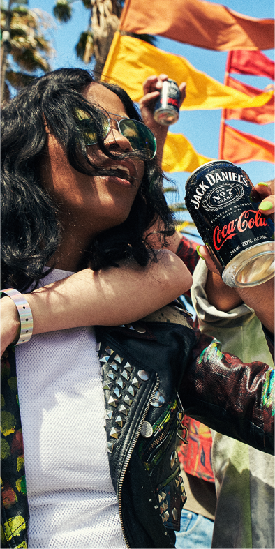 Outdoors image of an african-american woman in a leather jacket and aviator sunglasses holding a Jack Daniel's and Coca-Cola can with yellow and orange flags waving in the wind in the background. A man has his arm around her while they enjoy the Jack Daniel's and Coca-Cola beverage.