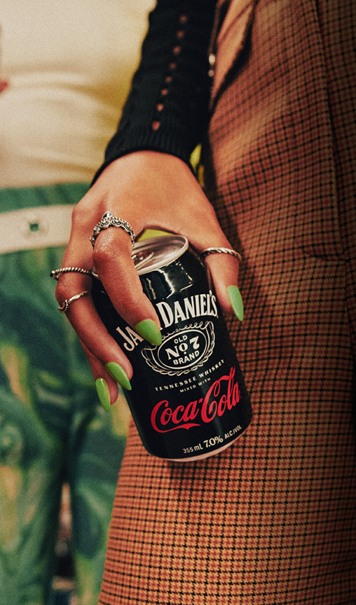 Hand with lime green manicured fingernails holding a Jack Daniel's and Coca-Cola can against orange plaid pants. In the background there is another woman wearing green and yellow swirled pants and a cream shirt holding another Jack Daniel's and Coca-Cola can.