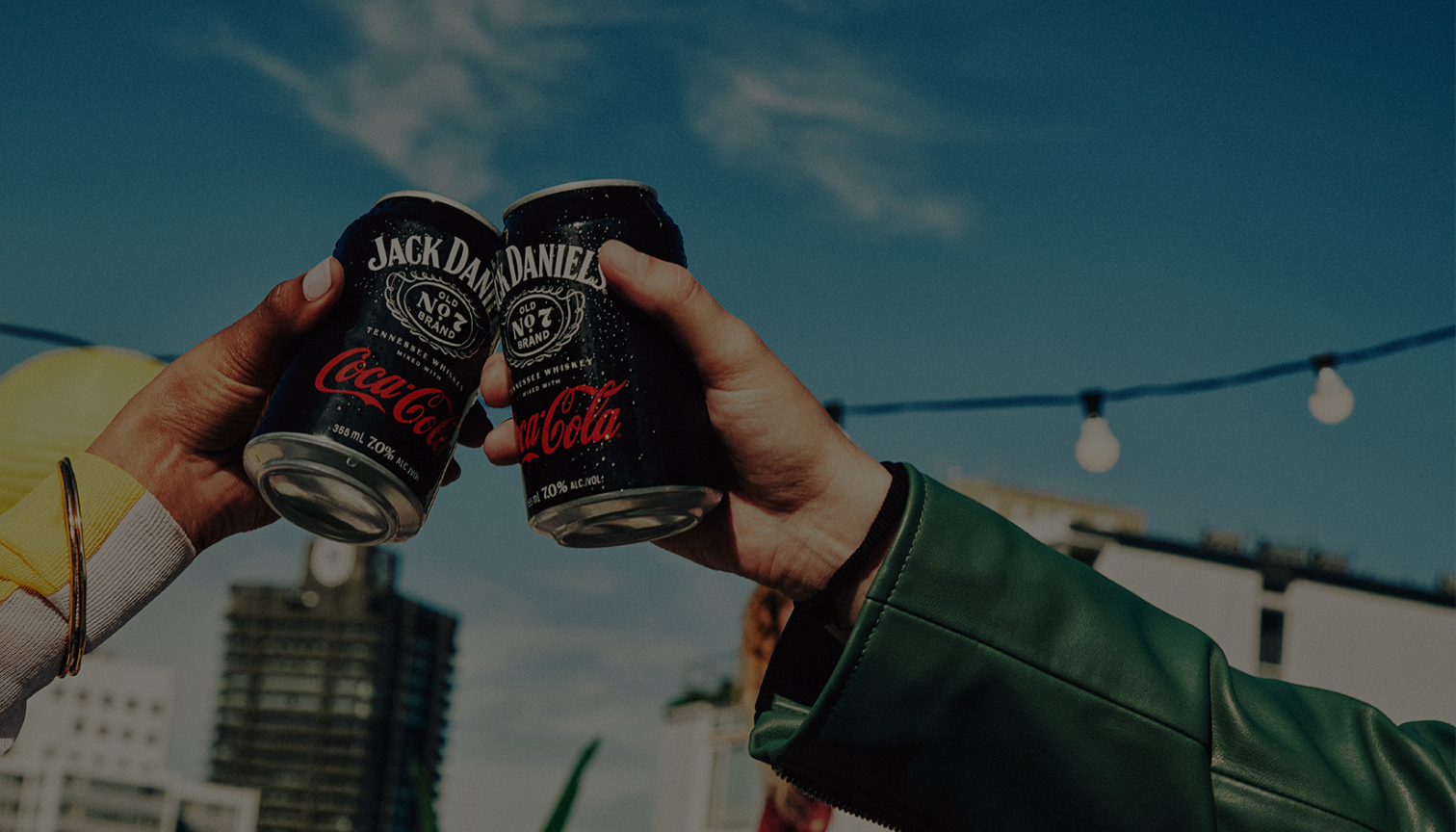 Image of an arm in a green jacket holding a Jack Daniel's and Coca-Cola can pressing up against another Jack Daniel's and Coca-Cola can held by an african-american arm in a cream and yellow sweater. Cheers!